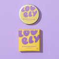 Banana Milkshake lip balm shown with its round tin lid and matching square box. The packaging features a bright yellow base with retro purple bubble lettering spelling 'LOVELY' and purple text identifying the flavor and product type.