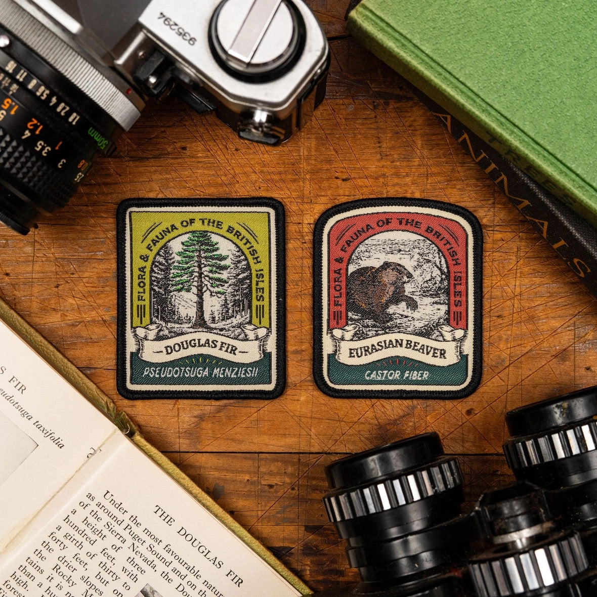 Two rectangular embroidered patches with rounded corners displayed on a wooden tabletop, surrounded by vintage camera lenses, an open book, and a green book. The left patch is labeled Douglas Fir and the right patch is labeled Eurasian Beaver, both with illustrated forest animals and trees.