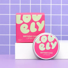 A pink tin of Lovely Skincare's Birthday Cake Lip Balm with its matching pink box, displayed on a white block against a purple tiled background.