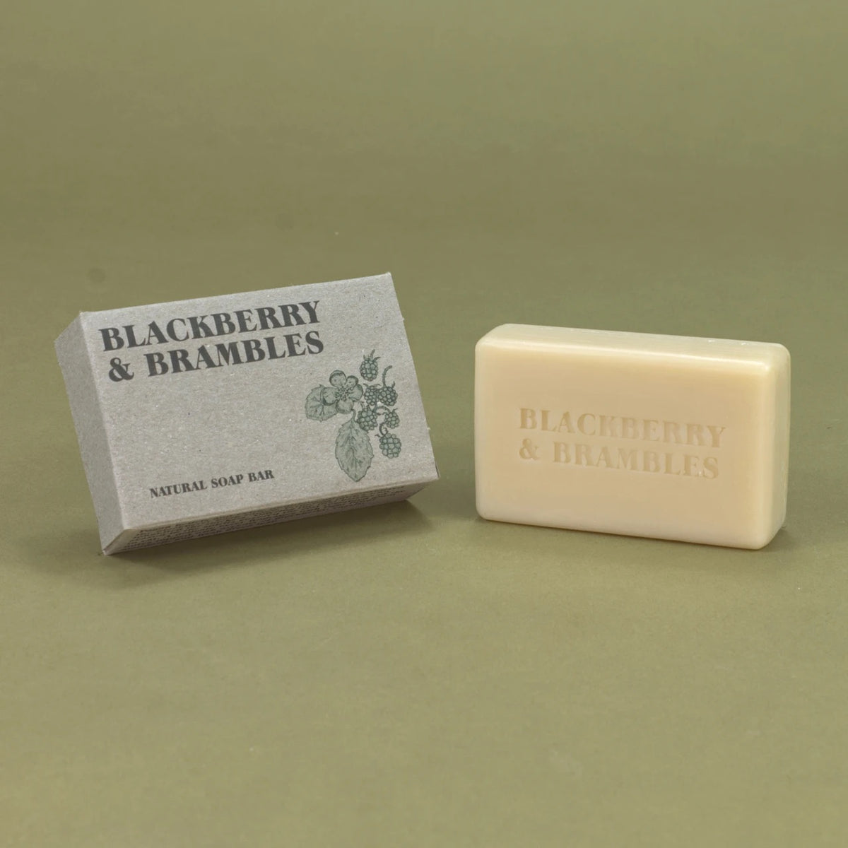 A bar of natural soap next to its textured gray packaging box, both embossed and printed with the brand name 'Blackberry & Brambles' on a muted olive green background.