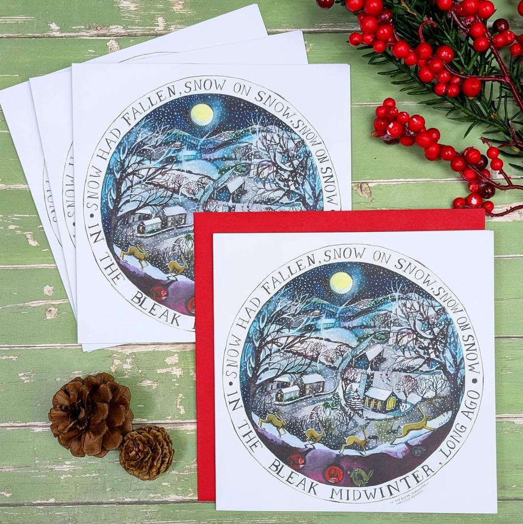 A Christmas card with a circular winter village illustration and red envelopes, arranged on a distressed light green wooden surface with pinecones and red berries as props.