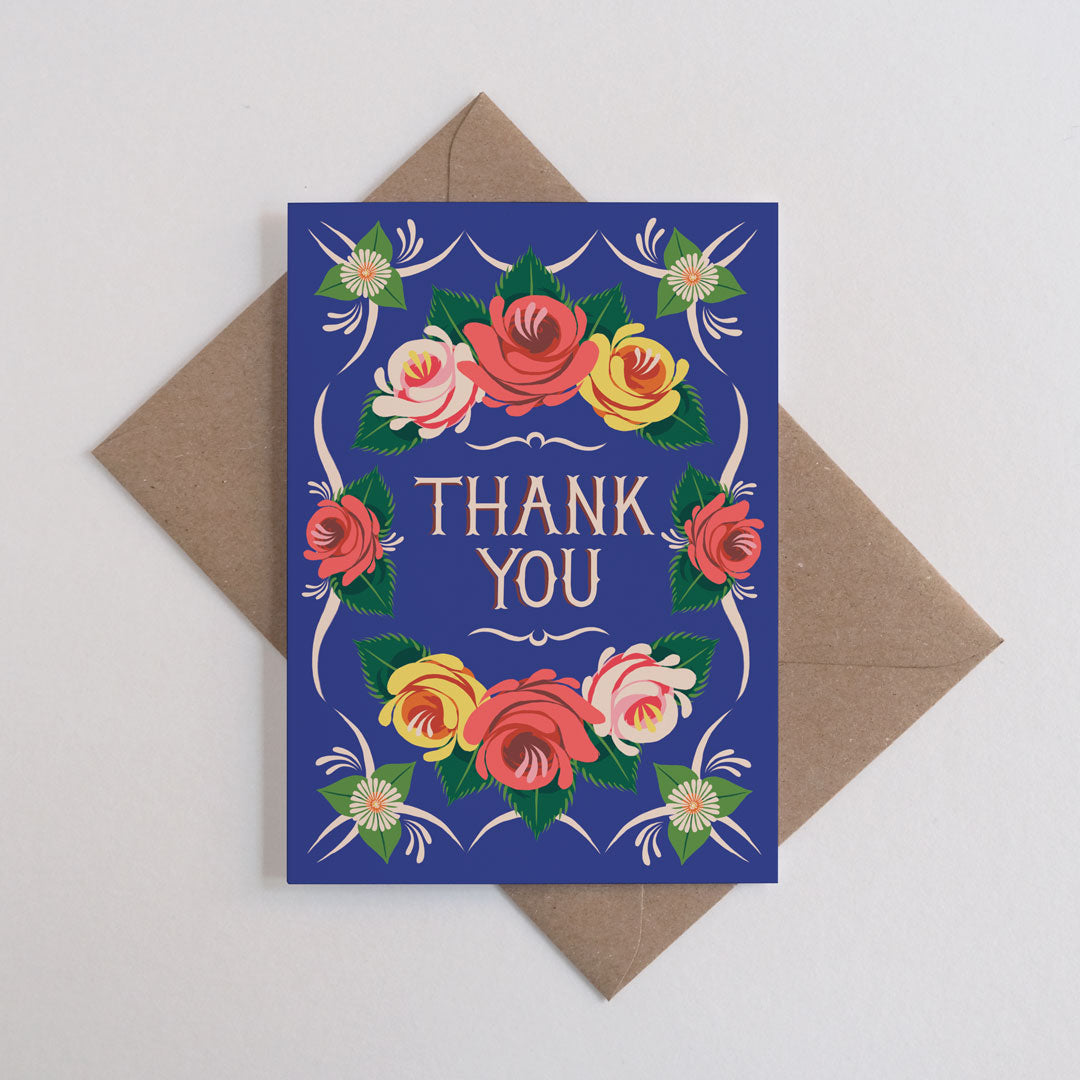 Rectangular thank you card with blue background and roses in yellow and pink, shown against a brown kraft envelope on a white backgrund.