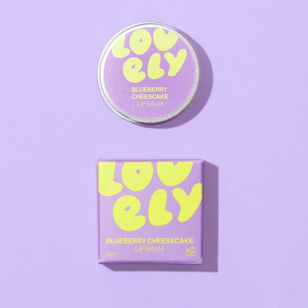 Lovely Skincare's Blueberry Cheesecake Lip Balm shown with its round tin and matching square box on a solid lavender background. The packaging features a light purple background with bold, playful yellow text spelling 'LOVELY' and smaller yellow text indicating the flavor.