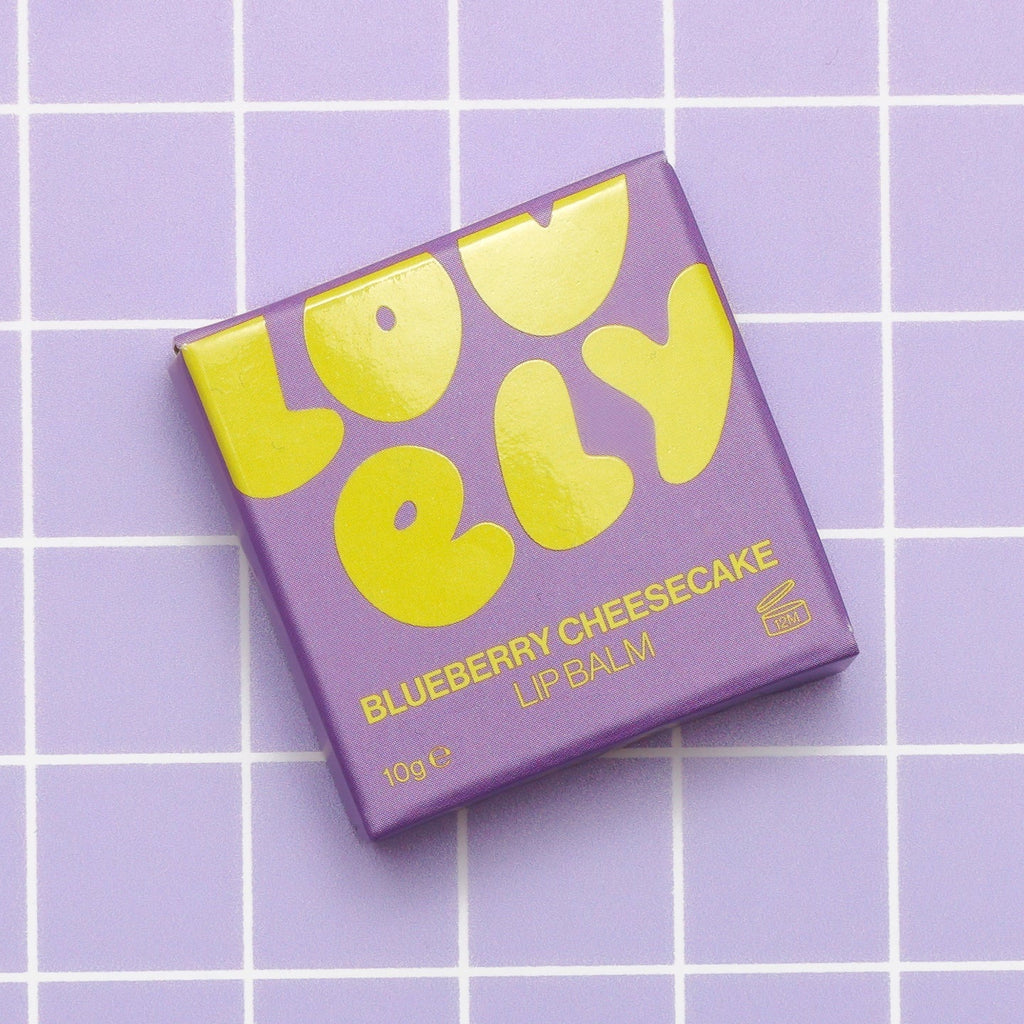 A square box of Lovely Skincare's Blueberry Cheesecake Lip Balm, featuring bold yellow lettering, placed on a light purple grid background.