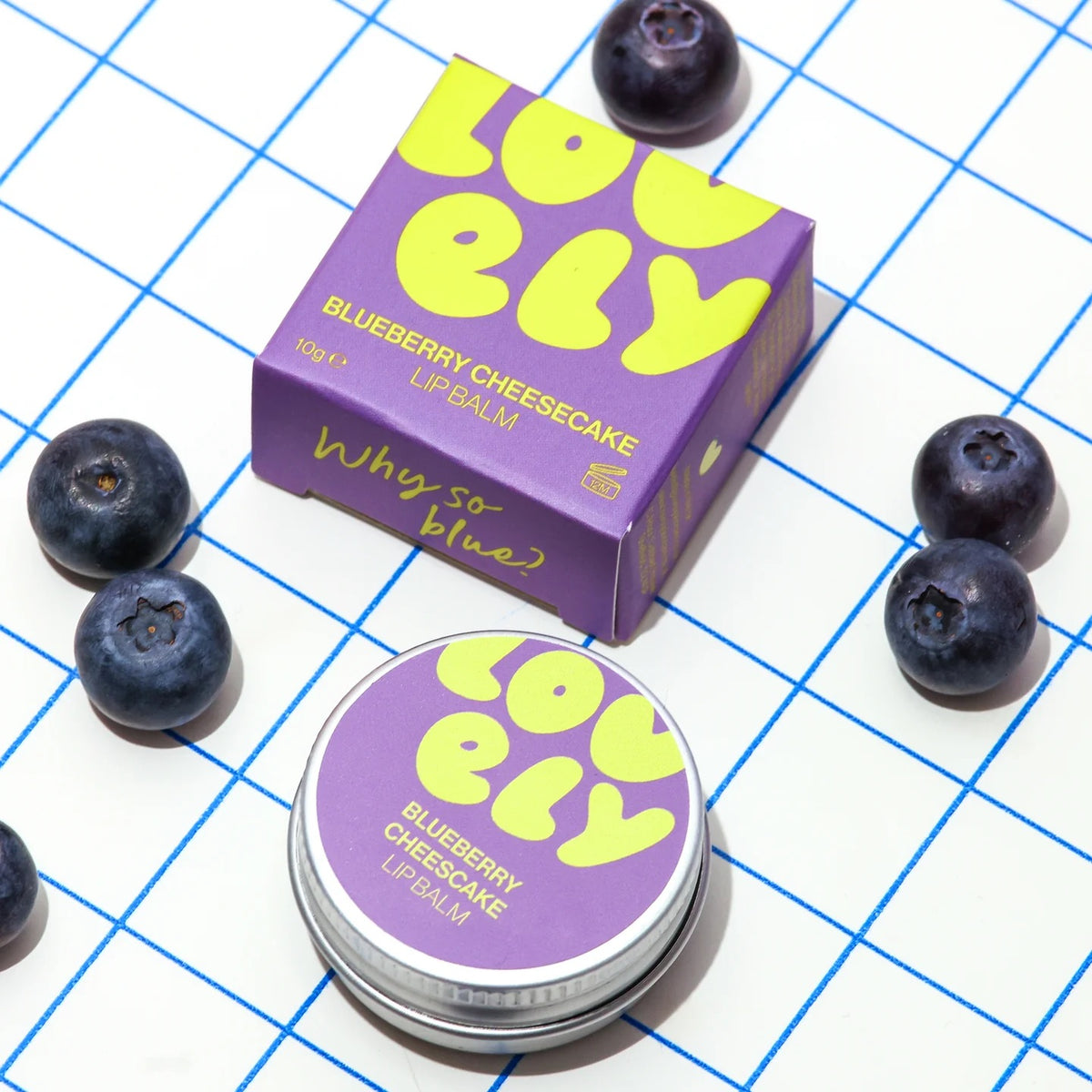 A box of Lovely Skincare's Blueberry Cheesecake Lip Balm arranged on a white surface with a blue grid pattern and scattered blueberries. The packaging is purple with bright yellow lettering.
