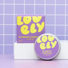 Blueberry Cheesecake flavoured lip balm in a round metal next to it's box on a purple background