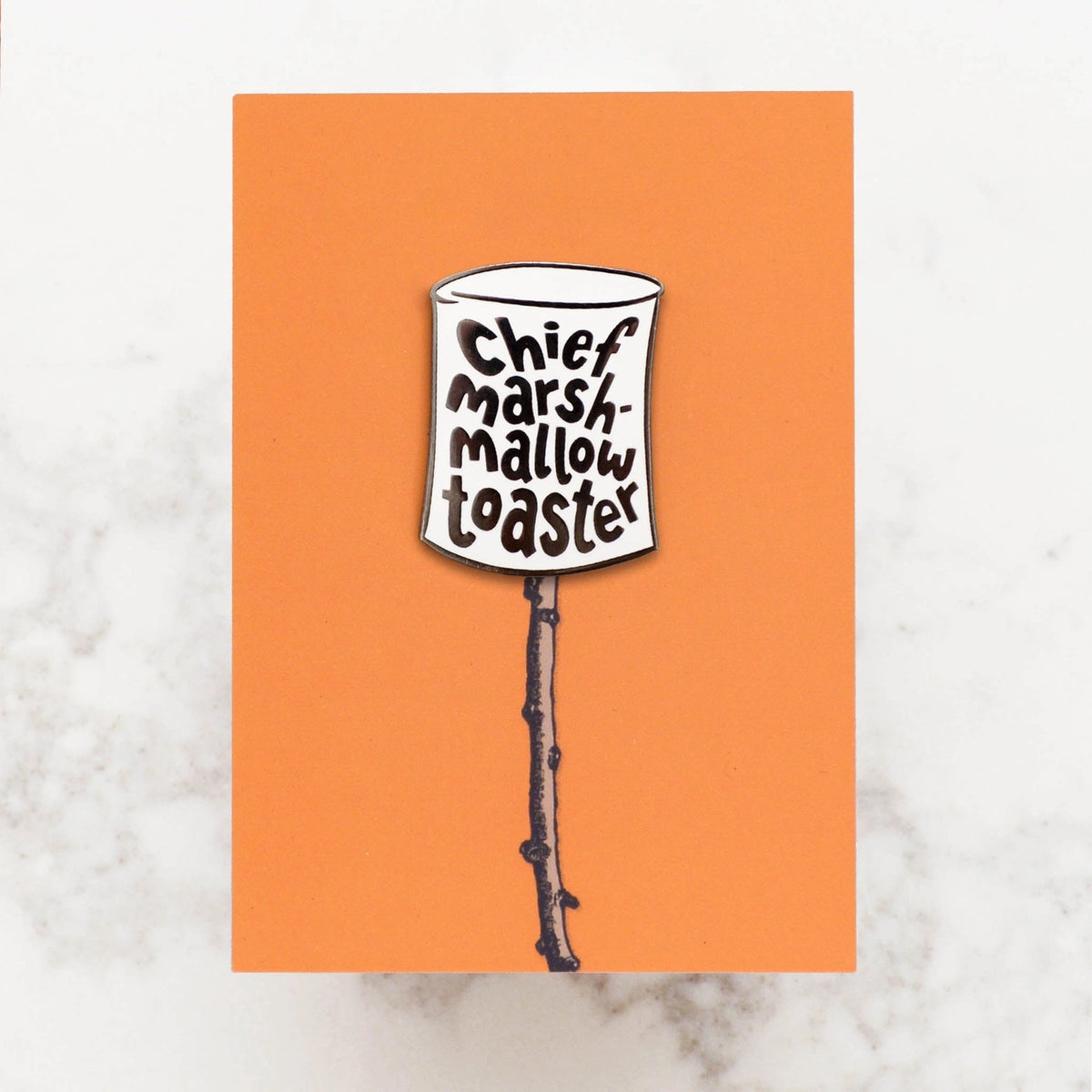 Enamel pin badge in shape of marshmallow with text reading 'Chief Marshmallow Toaster', displayed on an orange backing card with stick illustration.