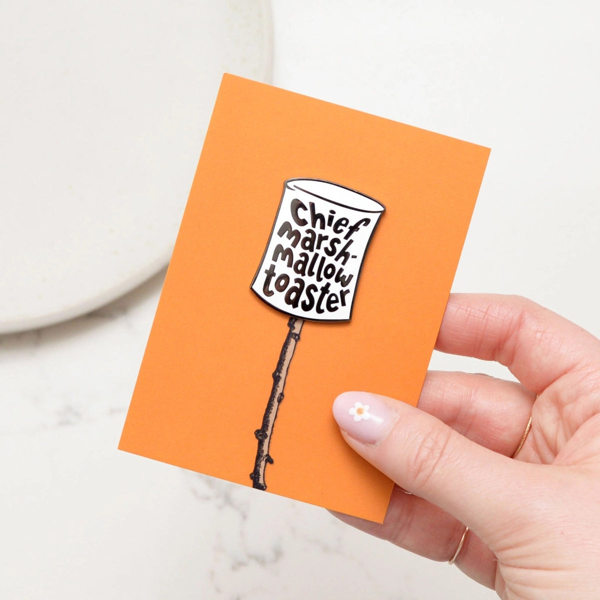 Hand holding card featuring Enamel pin in shape of marshmallow with text reading 'Chief Marshmallow Toaster'.