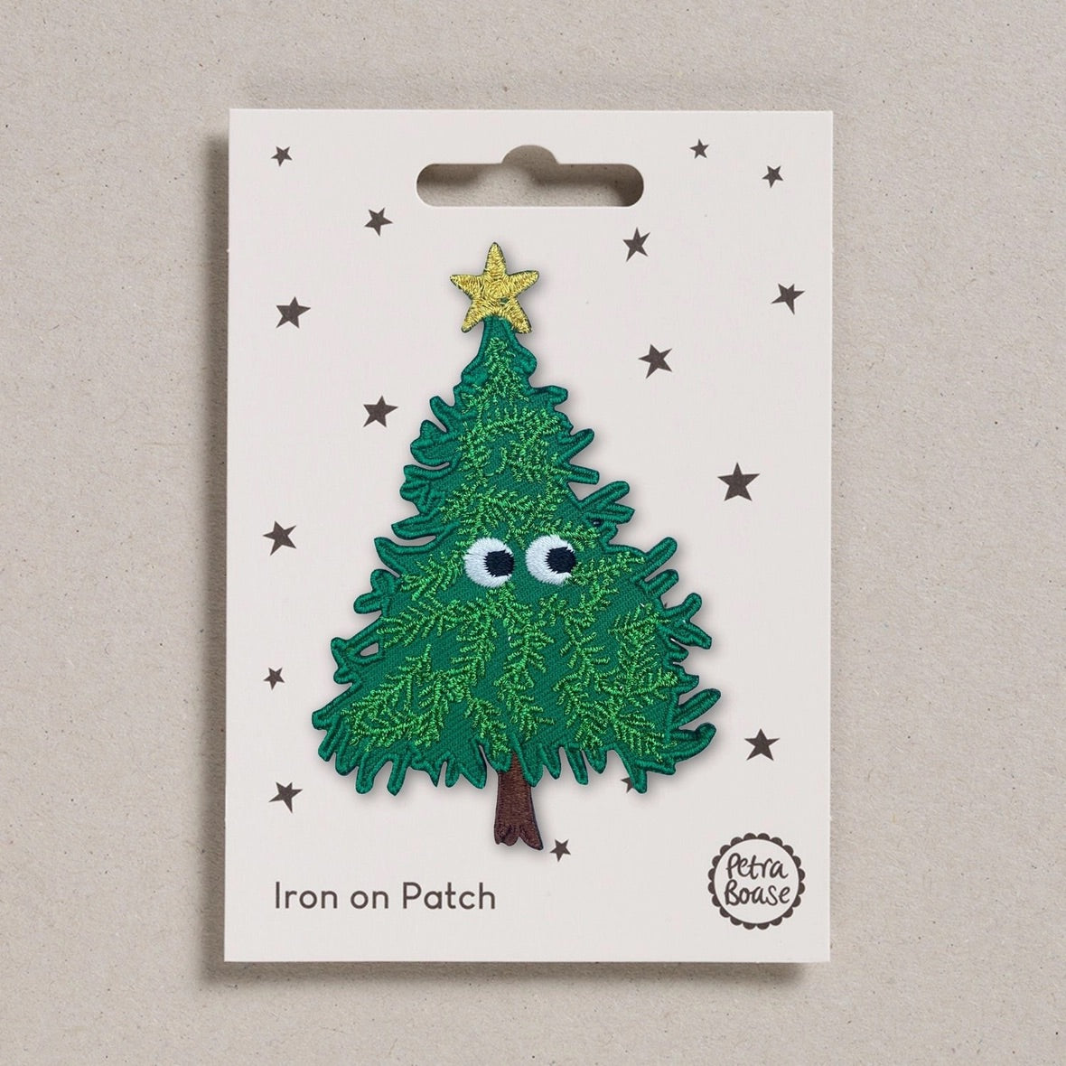 A Christmas tree iron-on patch with googly eyes and a gold star on a white backing card, photographed against a neutral beige background.