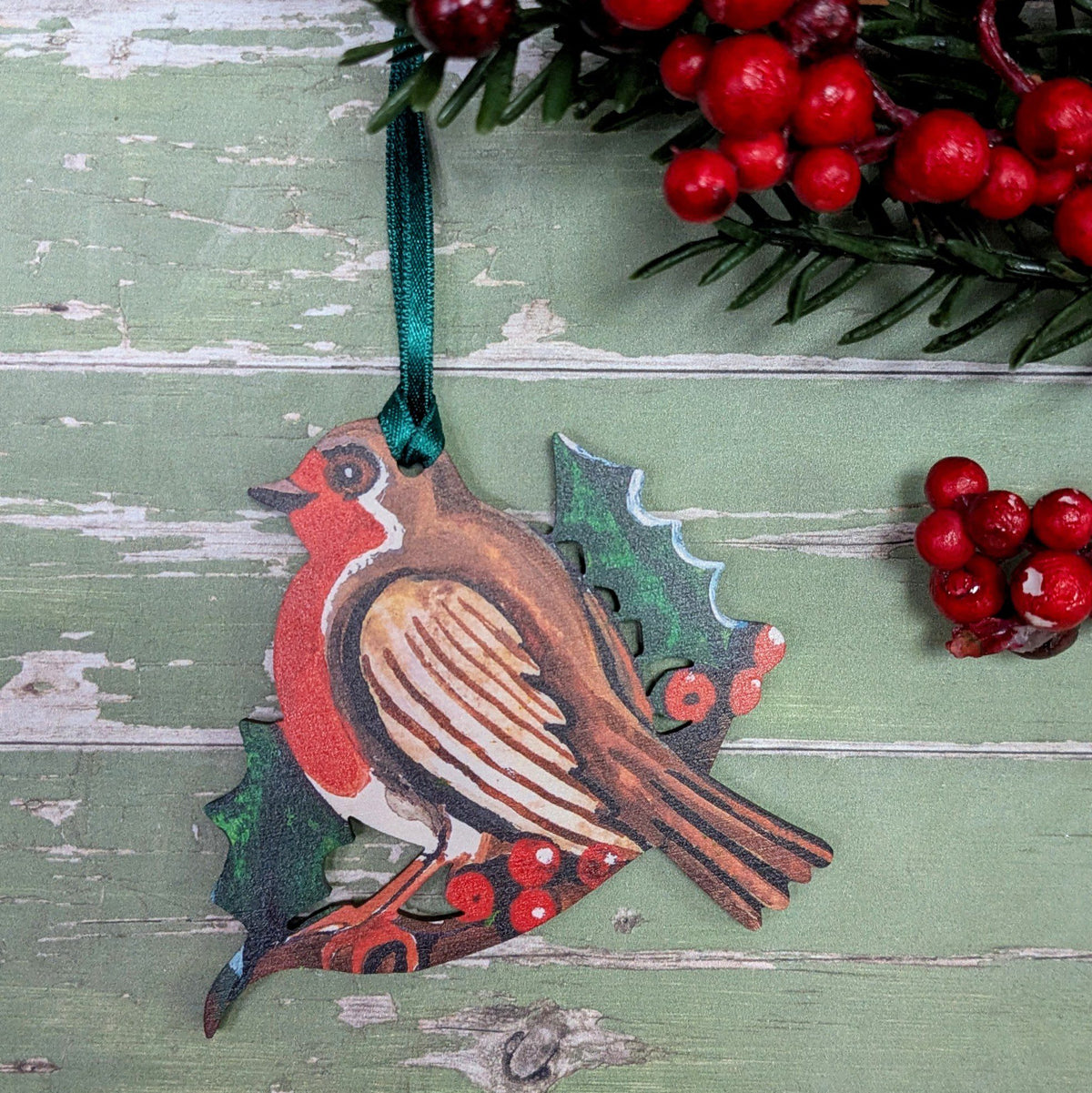 A Christmas inspired robin decoration made from wood. The robin is perched on a sprig of holly with it's dark green spiky leaves and vibrant red berries.