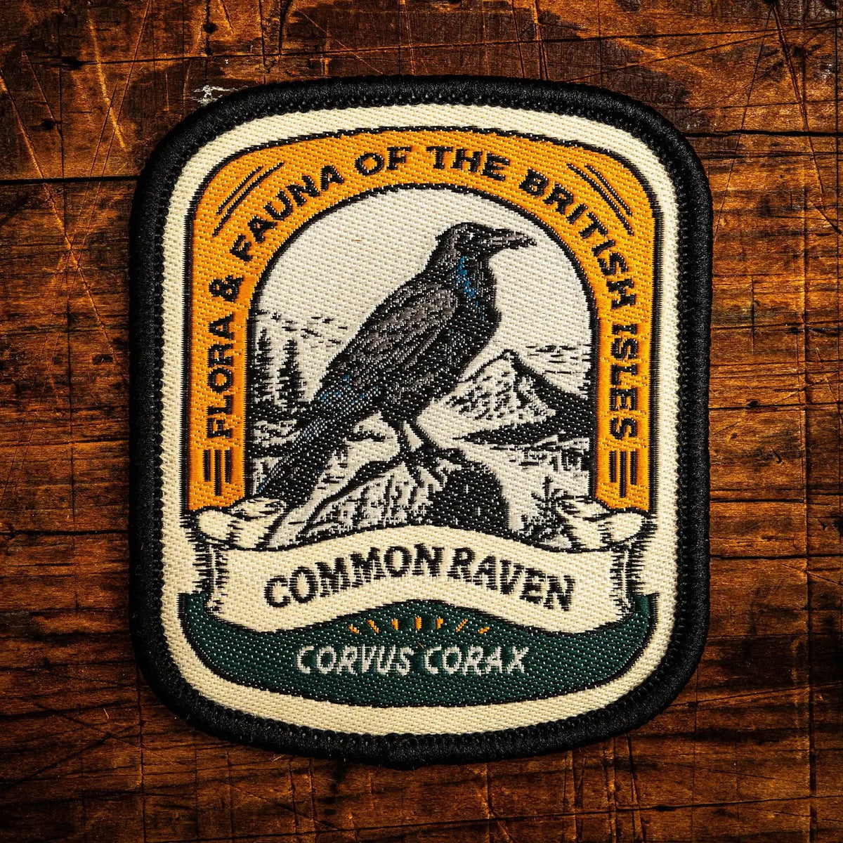 An embroidered patch featuring a black and white illustration of a common raven perched on a rock, with a yellow border reading 'FLORA & FAUNA OF THE BRITISH ISLES' and a banner reading 'COMMON RAVEN CORVUS CORAX', displayed on a rustic wooden background.