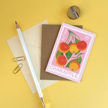 A flat-lay of a pink stamp-style mini card featuring autumn crab apples, placed on top of a brown envelope with a white pencil, paper clips, and a pencil sharpener on a yellow background.