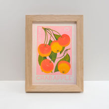 A framed art print featuring a vintage-style crab apple illustration with orange and yellow fruit, green leaves, and a pink background, set in a light natural wood frame against a neutral gray background.