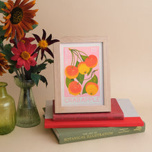 A framed art print featuring a vintage-style illustration of crabapples on a pink background, styled on top of stacked books next to a glass vase with flowers against a warm beige wall.
