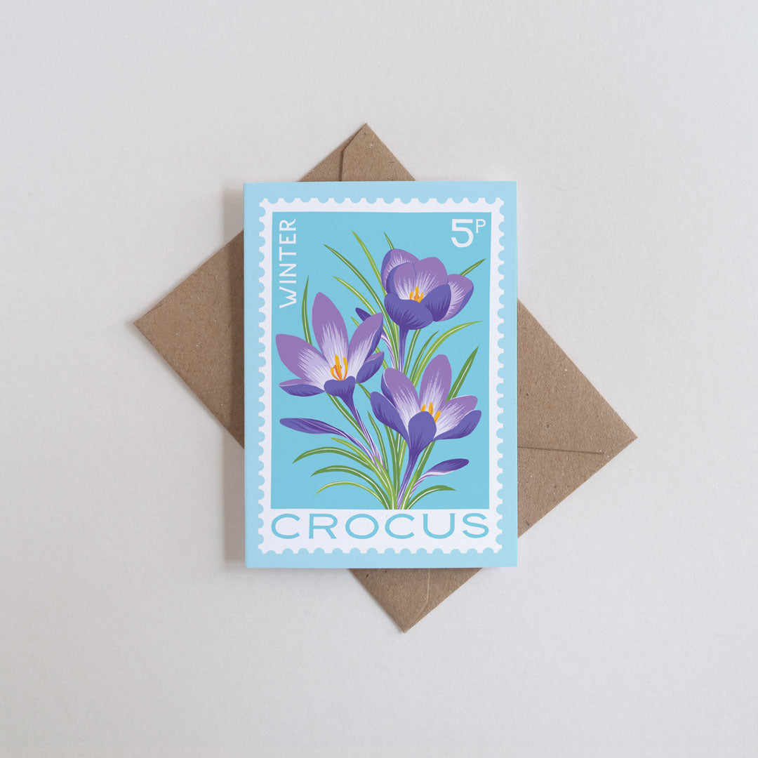 Rectangular mini card featuring a light blue background with an illustration of purple crocuses and green leaves, placed on top of a brown envelope against a light gray background.
