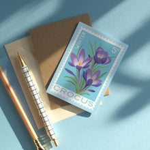 Greeting card with vintage-style postage stamp featuring purple crocuses on a light blue background. The card is styled on top of a brown envelope, with two gold pens on a light blue surface with soft shadows.
