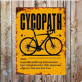 Yellow vintage style sign, showing signs of rust that has a picture of a push bike with the title 'CYCOPATH'. The dictionary style desciption says - 'noun - A person suffering from chronic bike riding disorder with abnormal urges to ride and feel free'.