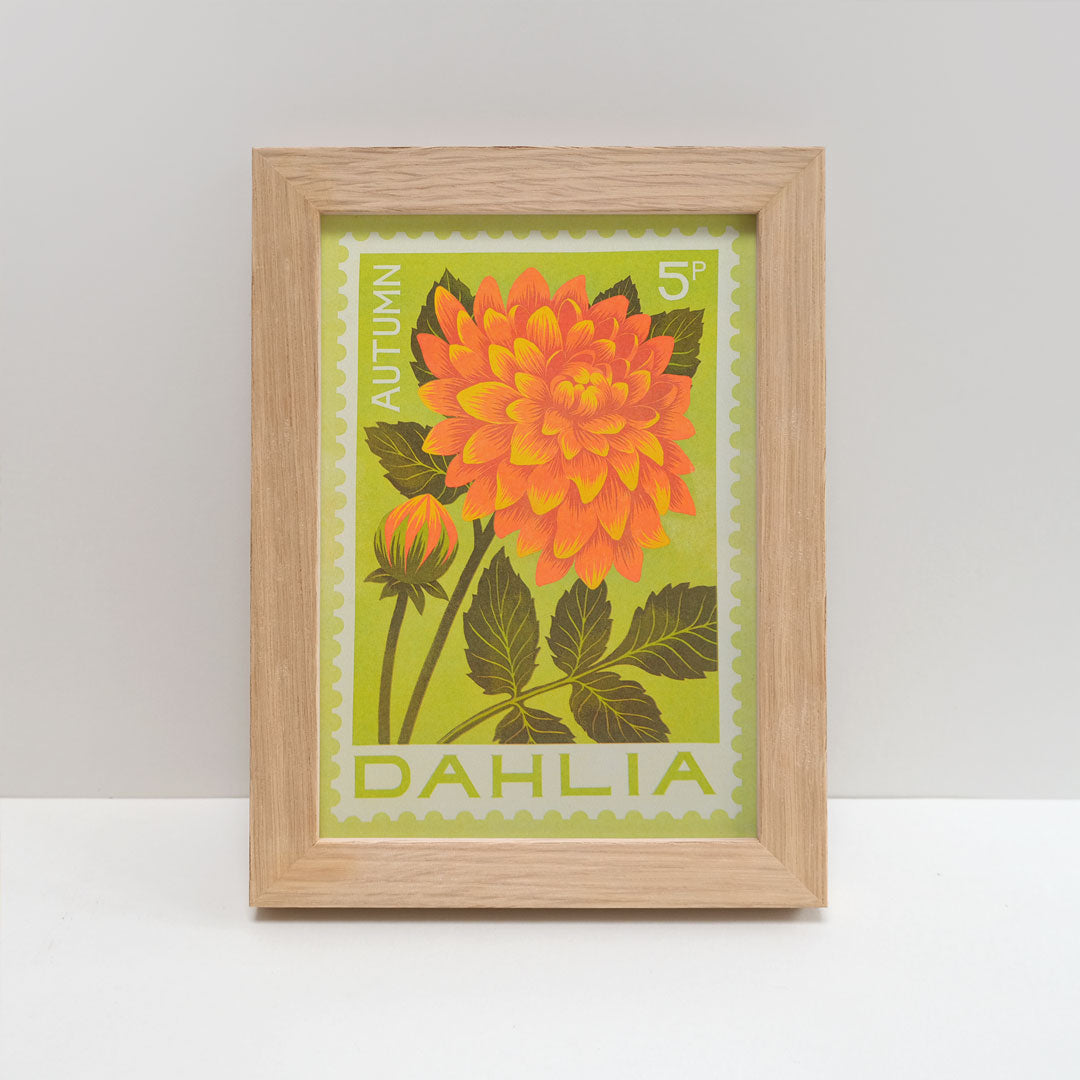 A light oak wood picture frame containing a vintage-style botanical art print of an orange autumn dahlia flower on a lime green background, displayed on a white surface against a light gray wall.