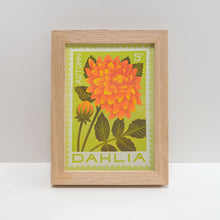 A light oak wood picture frame containing a vintage-style botanical art print of an orange autumn dahlia flower on a lime green background, displayed on a white surface against a light gray wall.