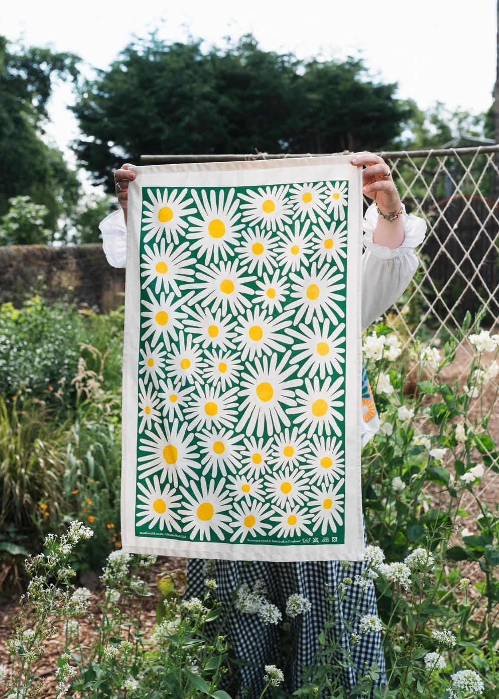 Daisy print tea towel on green background, displayed in a garden, held up by woman.