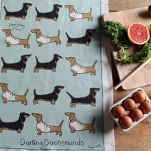 Tea towel with pale blue background and dachshund pattern with the text Darling Dachshunds at the bottom, next to a cutting board with eggs and vegetables