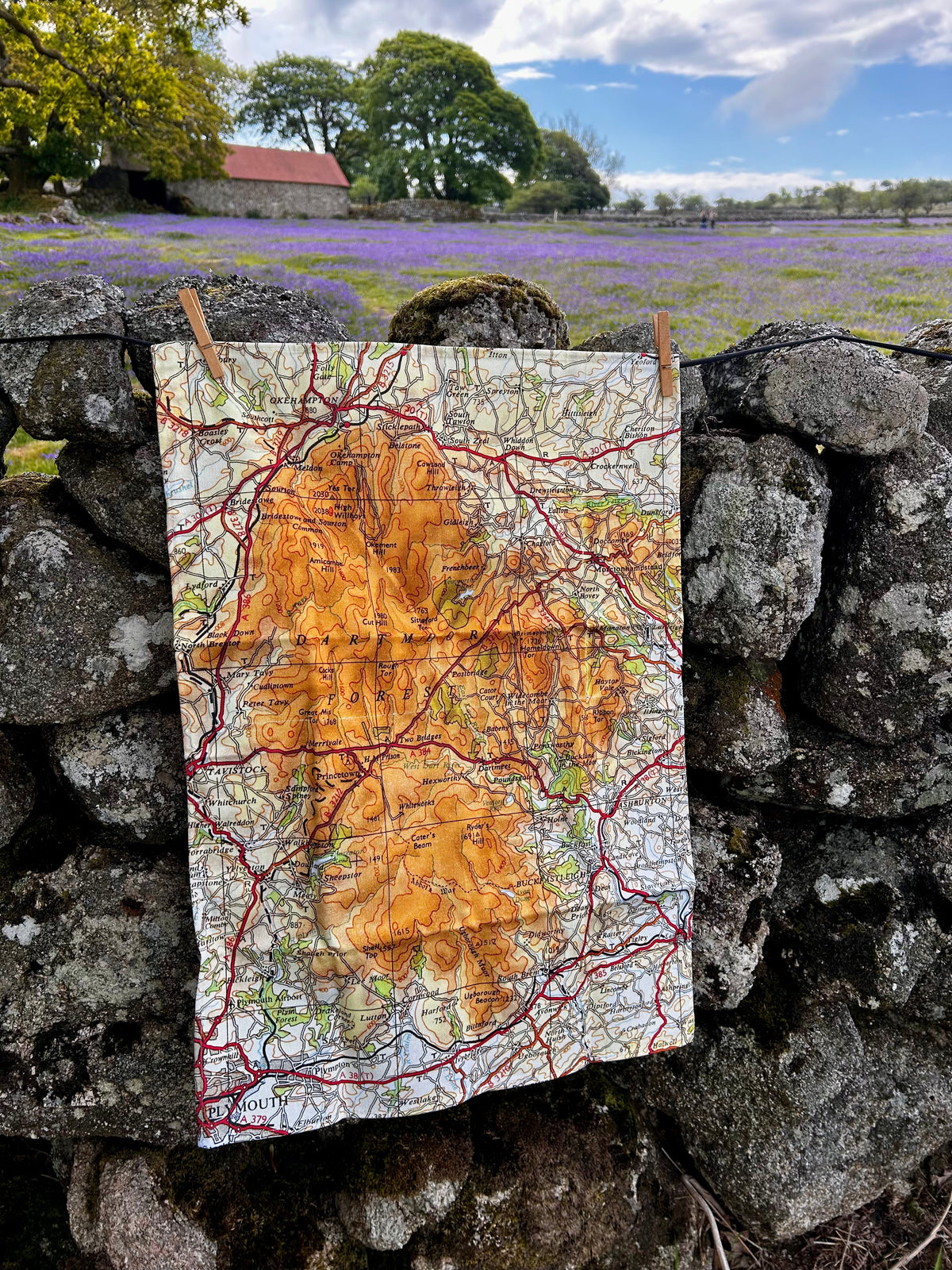 A Dartmoor map tea towel hanging from two wooden pegs on a rustic stone wall, with flowers, trees, and a barn visible in the background.