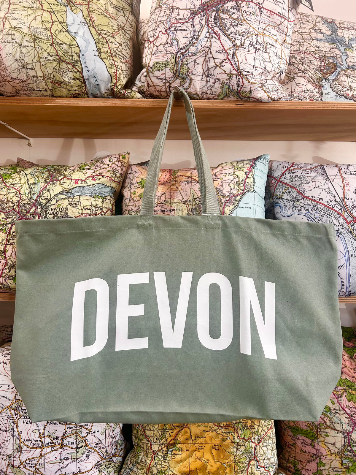 Green tote bag with 'DEVON' printed on it, hanging against a background of map-themed pillows.