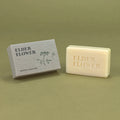 A cream-colored natural soap bar embossed with 'ELDER FLOWER' sits next to its textured gray packaging box on a solid olive green background.