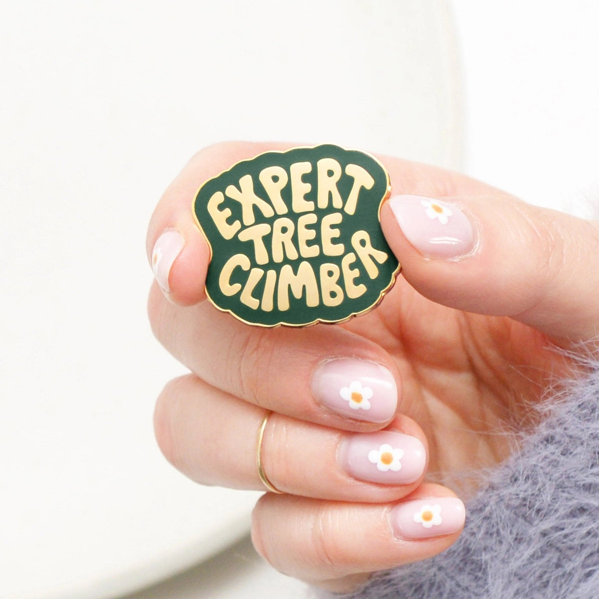 Hand holding a 'Expert Tree Climber' enamel pin with gold text and border and green background.