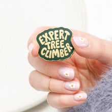 Hand holding a 'Expert Tree Climber' enamel pin with gold text and border and green background.