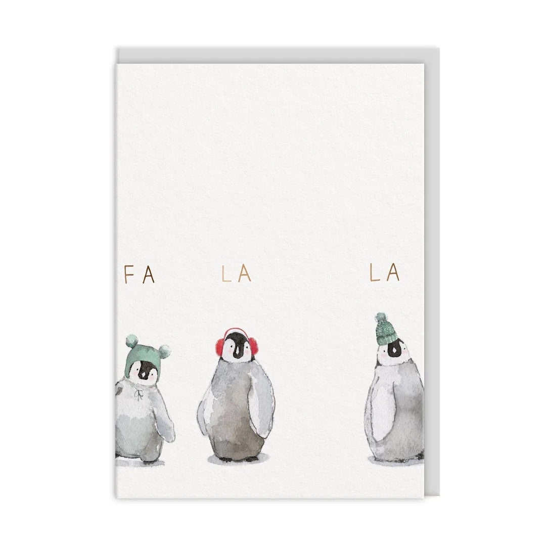 A Christmas card featuring three watercolor penguin illustrations wearing green and red winter accessories, with the letters 'FA LA LA' printed above them. A grey envelope is partially visible behind the card on a white background.