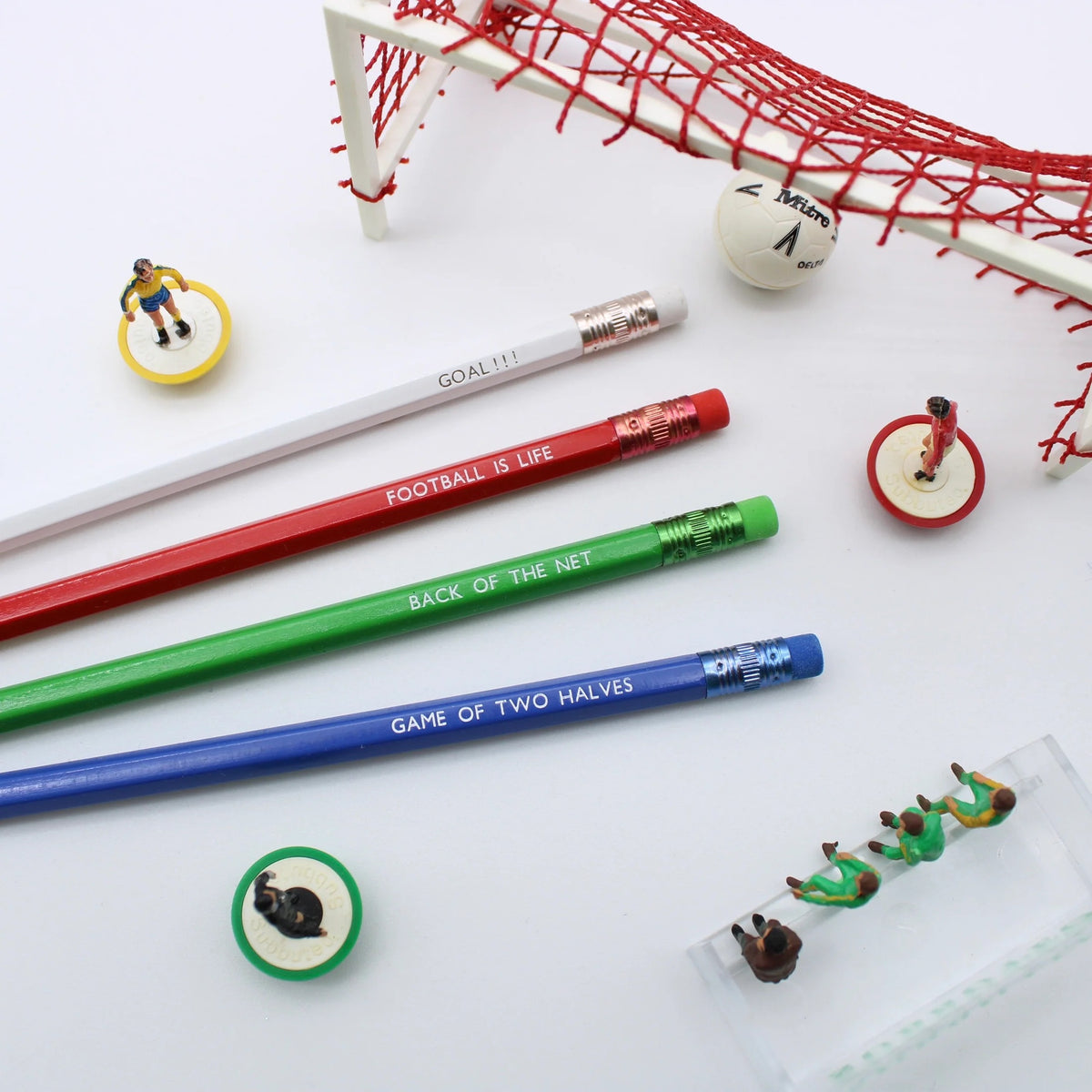 Four soccer-themed pencils in white, red, green, and blue arranged on a white background, surrounded by miniature soccer players including a small ball and a goal net.