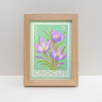A framed art print featuring a vintage-style crocus stamp illustration with three purple flowers on a mint green background, set in a light natural wood frame.