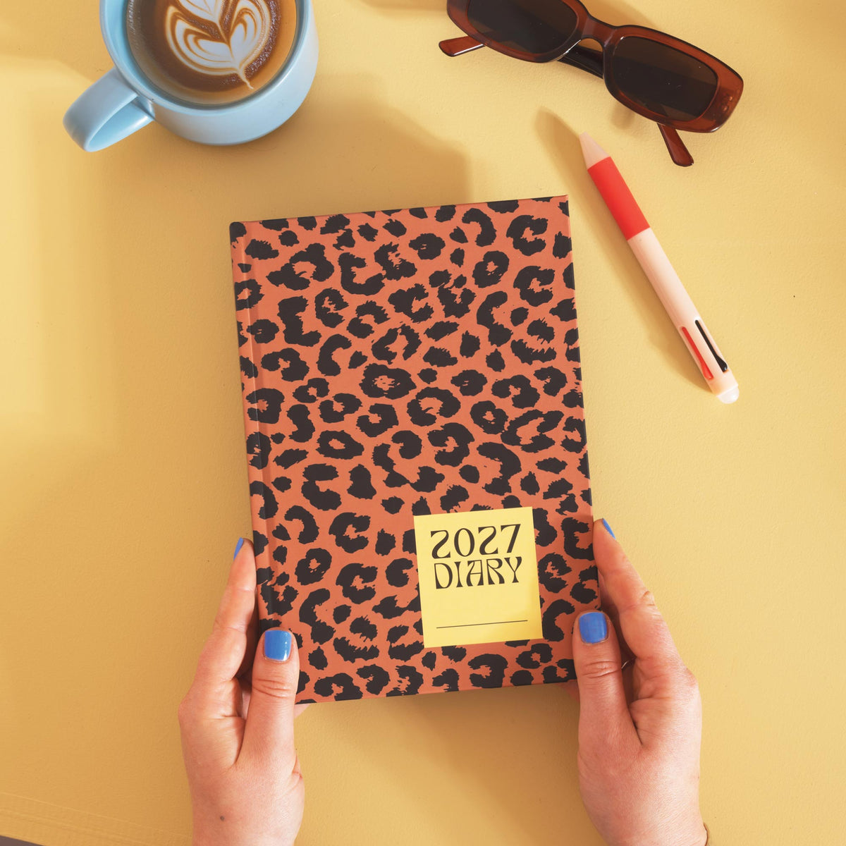 Leopard print diary with '2027 Diary' label held by hands on a yellow background with a cup of coffee, sunglasses, and pen.