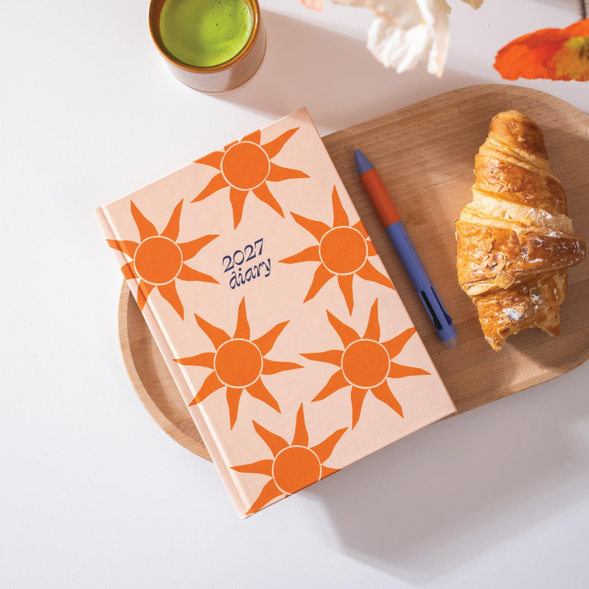 2027 diary with sun pattern, croissant, pen, and cup on a wooden board