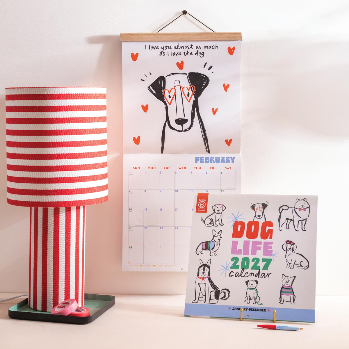  'Dog Life 2027' calendar on stand with open calendar hanging on wall on a light background with red and white striped lamp beside it.
