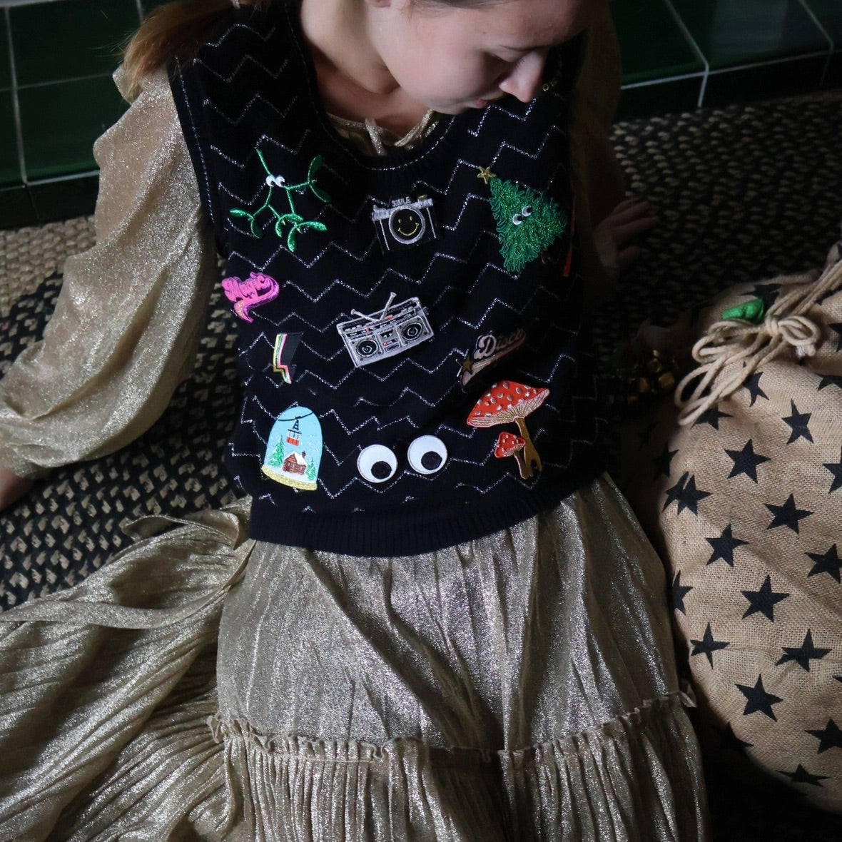 Child wearing a dark sweater with colorful patches sitting on a patterned surface.
