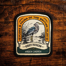A rectangular embroidered patch with rounded corners featuring a grey heron illustration and the text 'FLORA & PAUNA OF THE BRITISH ISLES', 'GREY HERON', and 'ARDEA CINEREA', placed on a rustic wooden surface.