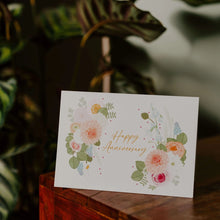 Greeting card with floral design and 'Happy Anniversary' text on a wooden surface with a blurred natural background.