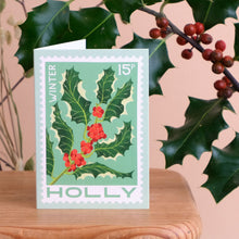 A vintage-style winter holly greeting card standing on a wooden surface, with a peach-colored background and real holly leaves and berries in the frame.