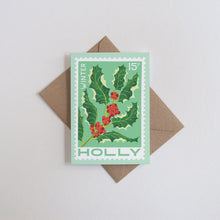 A vintage stamp style greeting card, featuring green holly leaves and red berries on a mint green background, placed on top of a brown kraft envelope against a light gray background.
