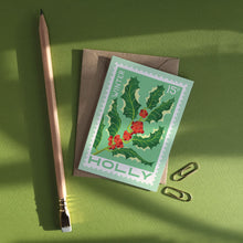 VIntage stamp style card with green holly and red berries against a mint green background. Displayed on a green table with stationery accessories.