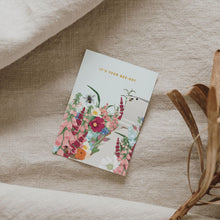 A birthday card with a floral bee-themed design lies on a textured beige linen surface, styled with dried branches and soft fabric folds.