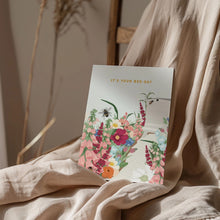 A floral greeting card with the text 'IT'S YOUR BEE-DAY' resting on draped beige linen fabric.