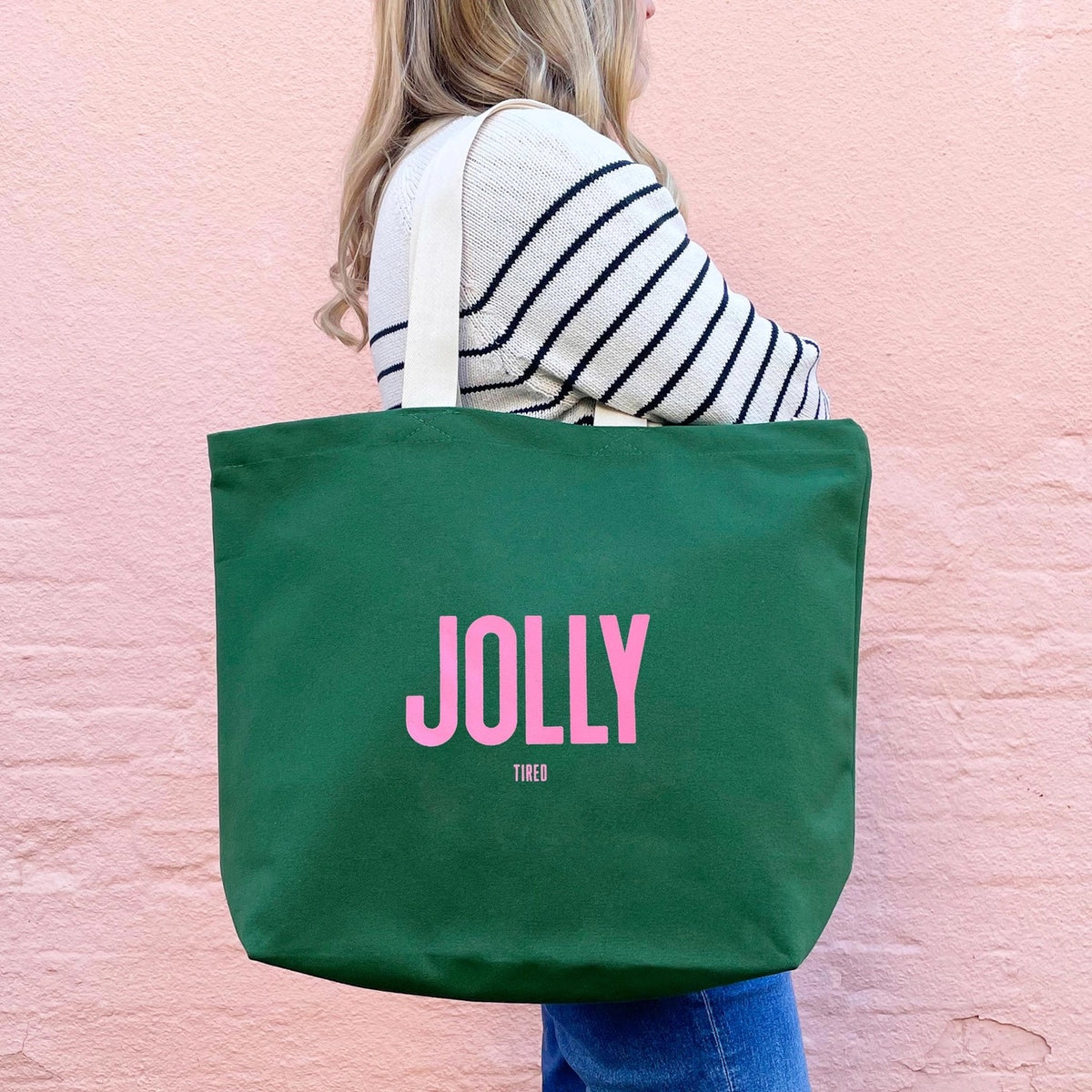Person holding a green tote bag with 'JOLLY TIRED' printed on it against a pink background