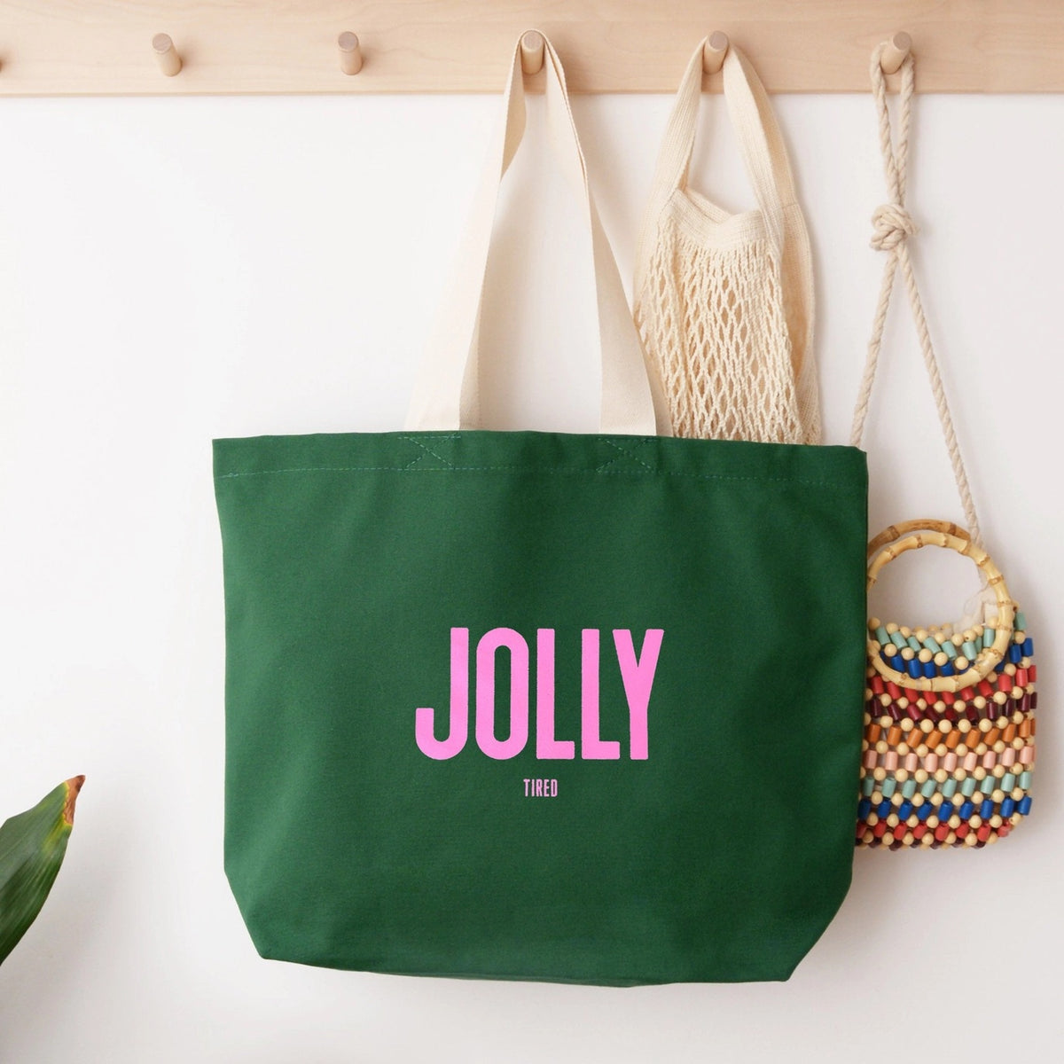 Green tote bag with 'Jolly Tired' text, hanging against a wall