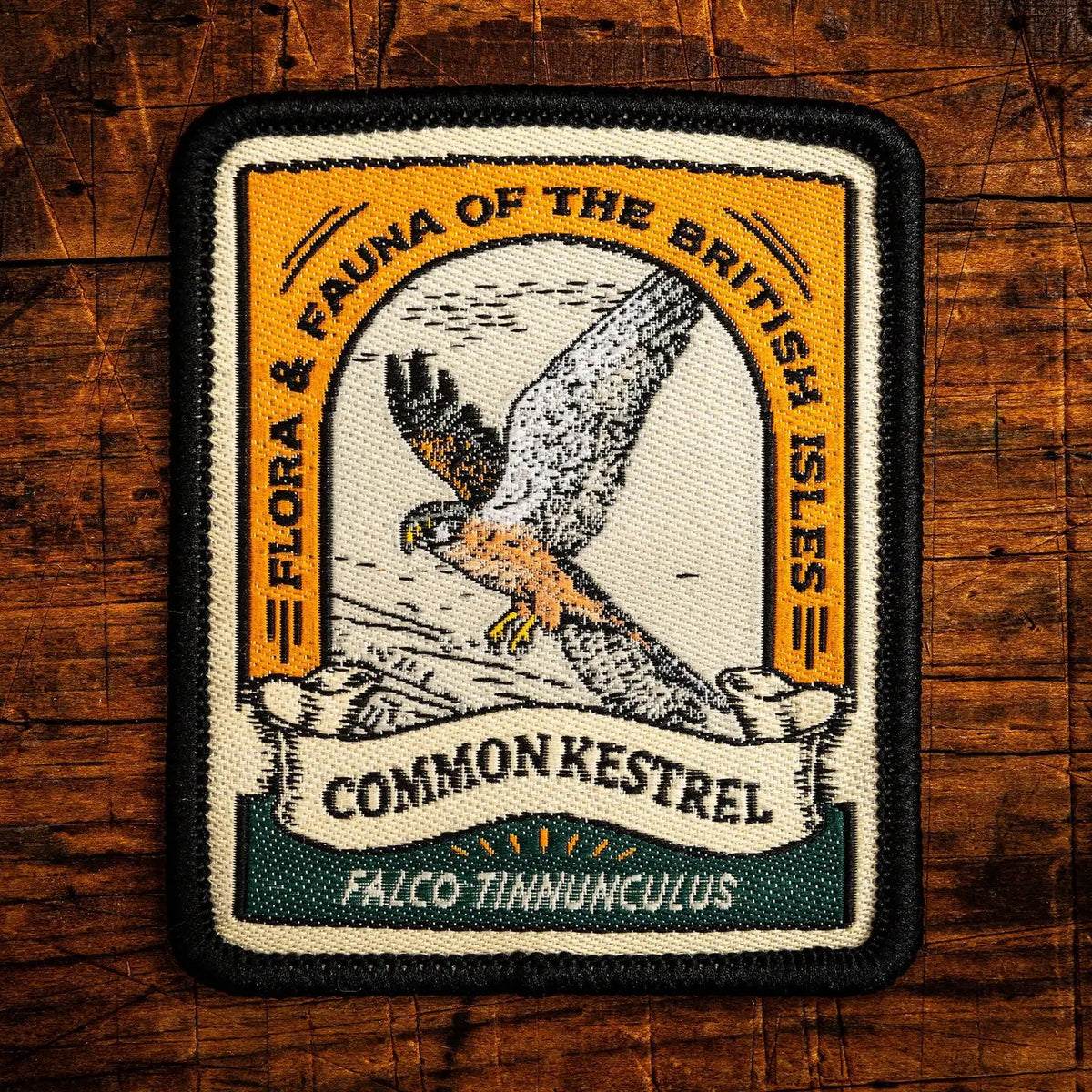 A rectangular embroidered patch with a black border and a vintage-style illustration of a kestrel bird, reading “Common Kestrel” and “Flora & Fauna of the British Isles,” placed on a rustic wooden surface.