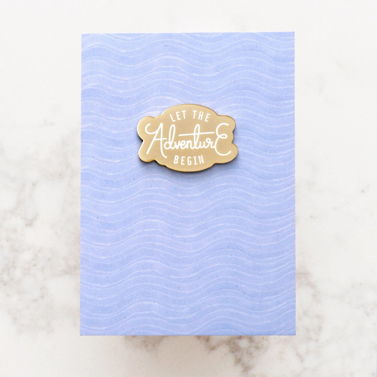 Gold shaped pin with white text 'Let the adventure begin' by Alphabet Bags, shown on blue backing card