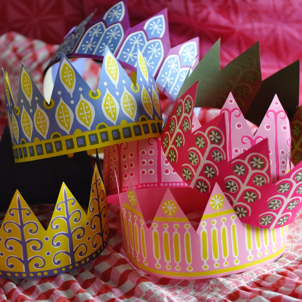 Three decorative paper crowns with colourful printed patterns, displayed on a red-and-white checkered fabric surface. These crowns are included in the carcker packs.