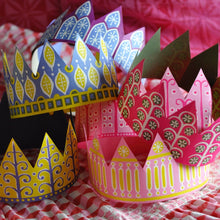 Three decorative paper crowns with colourful printed patterns, displayed on a red-and-white checkered fabric surface. These crowns are included in the carcker packs.